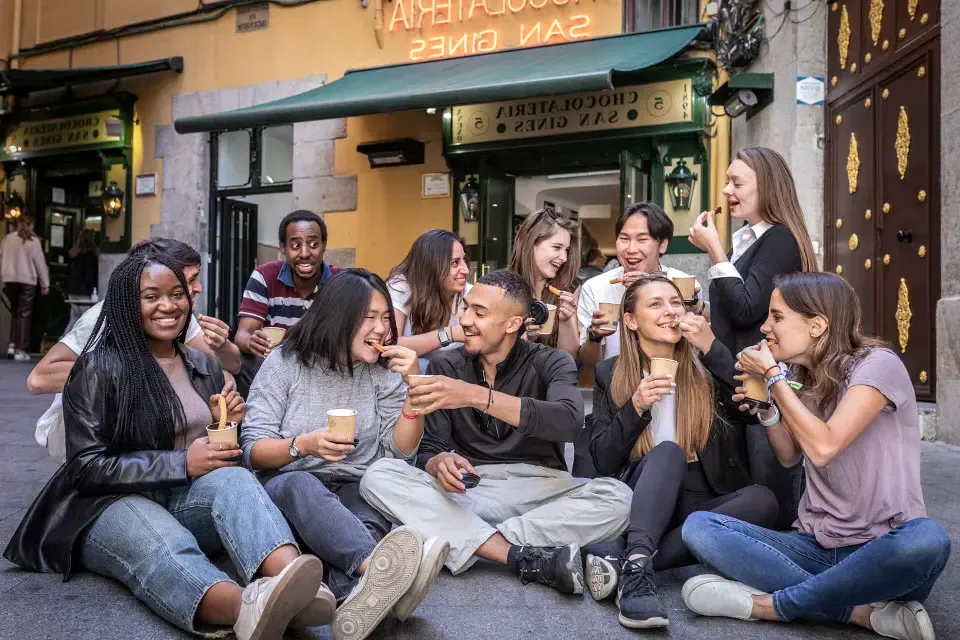 学生们坐在一家名为Chocolateria San Gines的商店前，把糕点泡在热巧克力杯里吃.